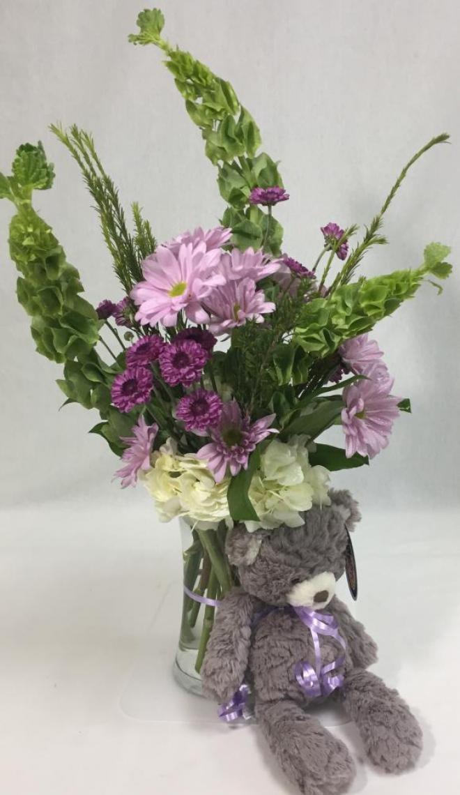 Purple chrysanthemum daisies, white hydrangeas, purple button mum daisies, bells of Ireland, and honey bracelet in a tall hour glass vase adored with a gray bear